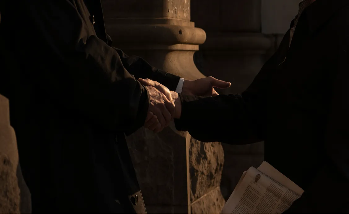 Two people shaking hands in front of a stone wall, one holding folded newspapers.