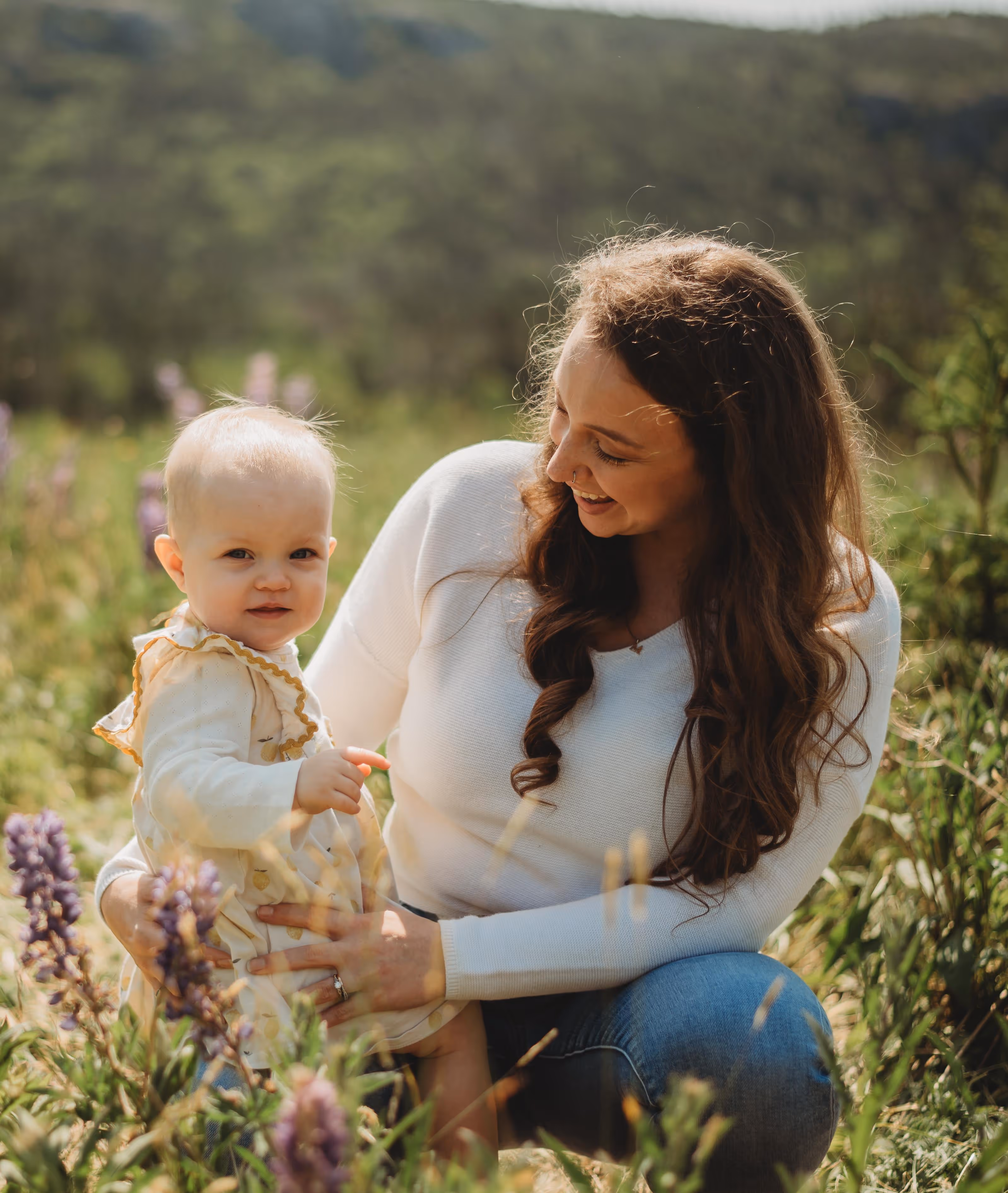 Woman with long curly hair in a white sweater holding a baby in a field with purple flowers.