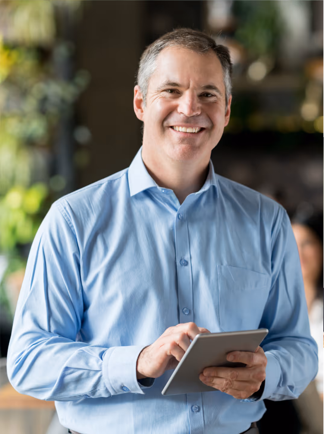Smiling man in a light blue shirt holding and using a tablet indoors with plants blurred in the background.