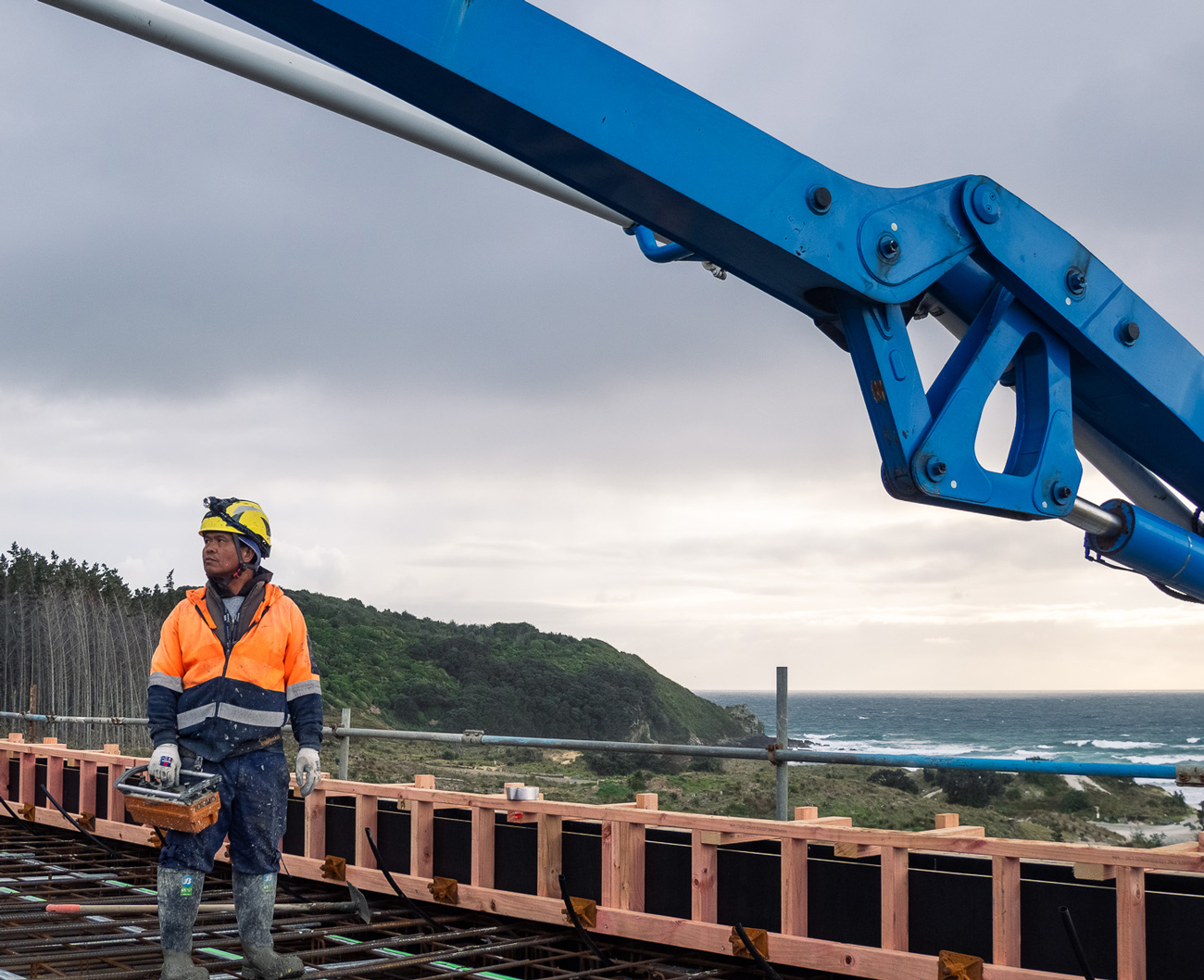 Construction worker in high-visibility gear operating concrete pumping equipment on a coastal building site in New Zealand.