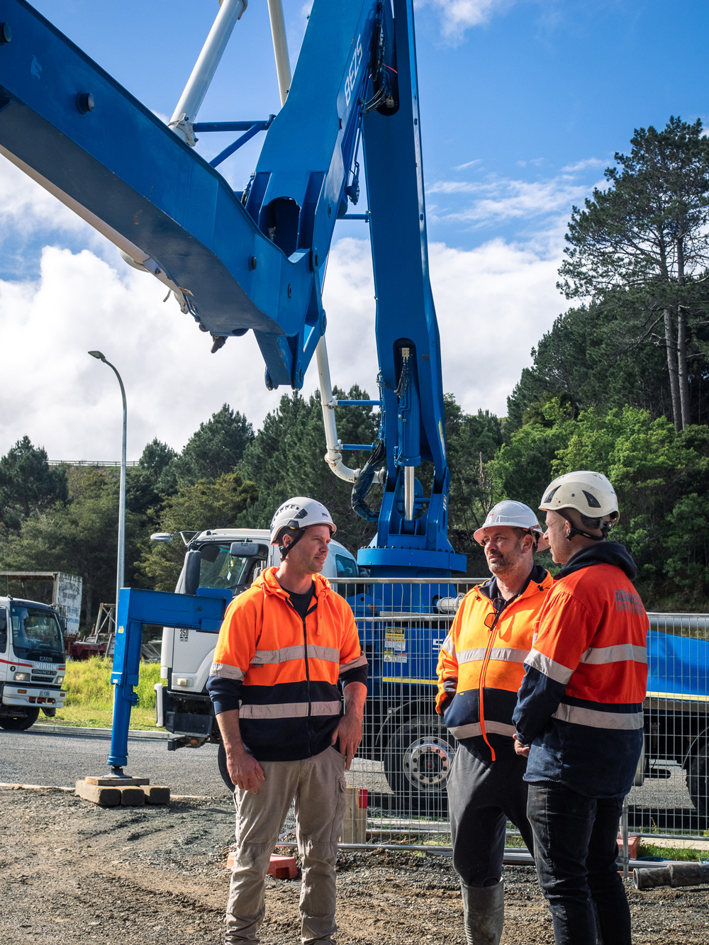 Three construction workers in orange high-visibility jackets and helmets standing beside a large blue concrete pump truck on a worksite."