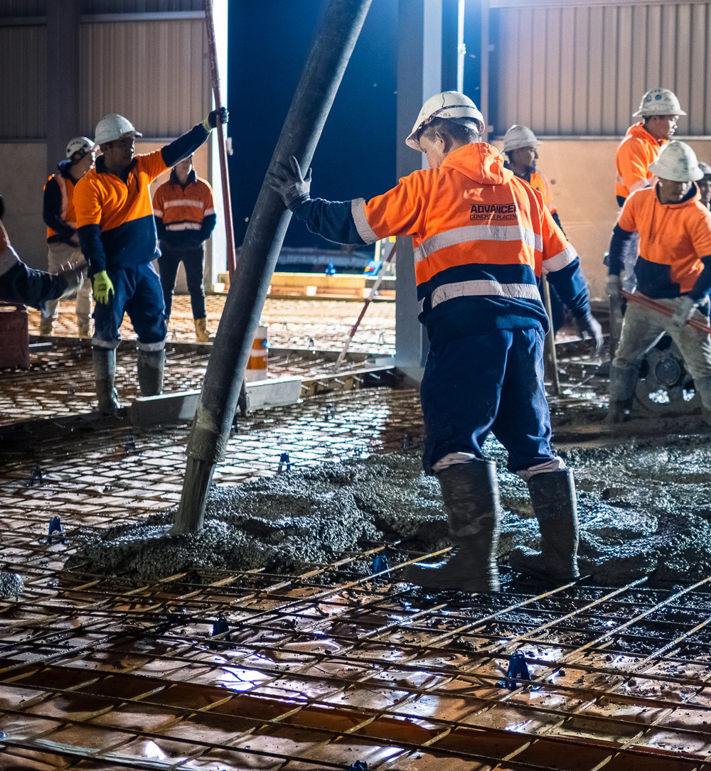 Construction crew in orange high-visibility jackets and helmets pouring and spreading concrete over reinforced steel mesh on a building site.