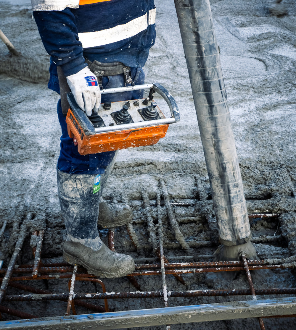 Construction worker in safety gear operating a remote control while concrete is poured over steel reinforcement on a building site.