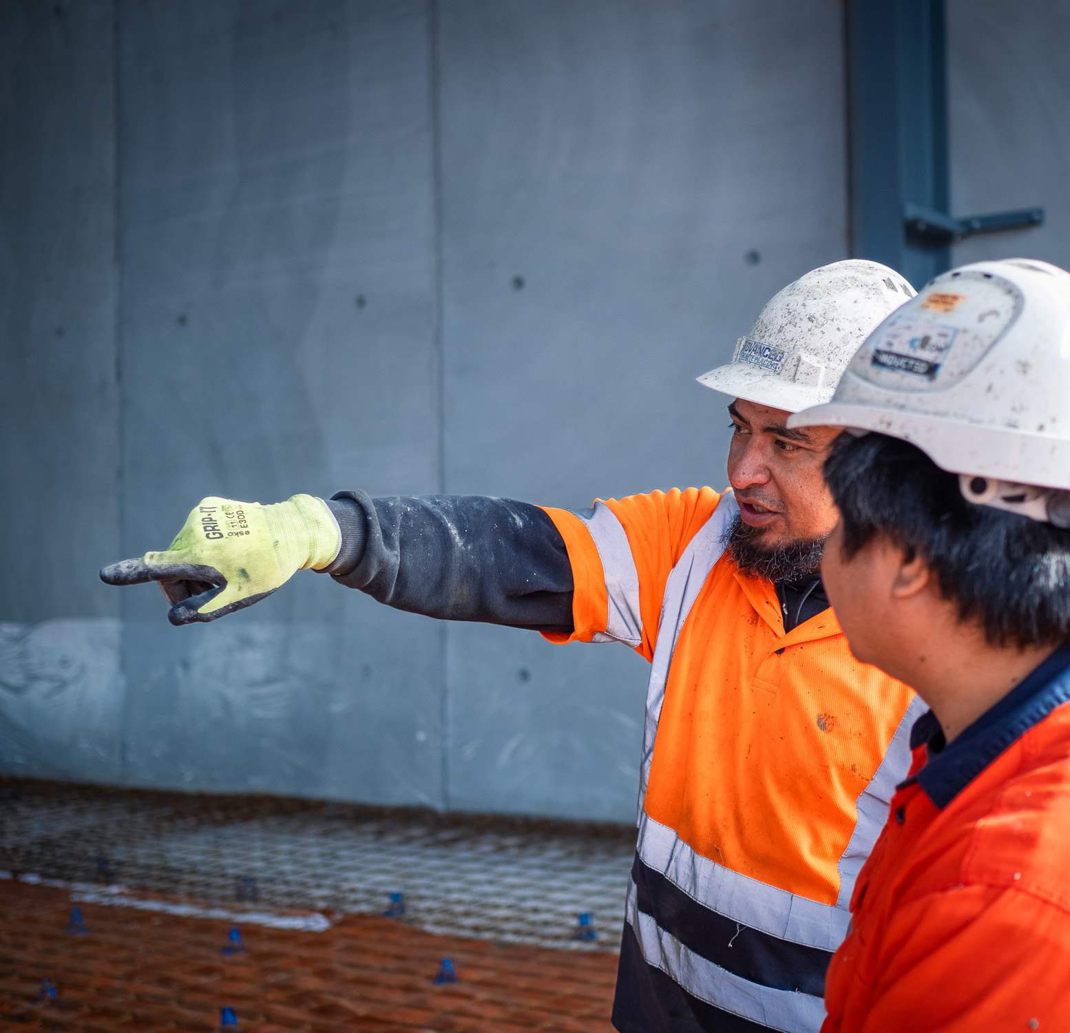 Construction worker in high-visibility gear pointing while talking with a colleague on a concrete worksite.