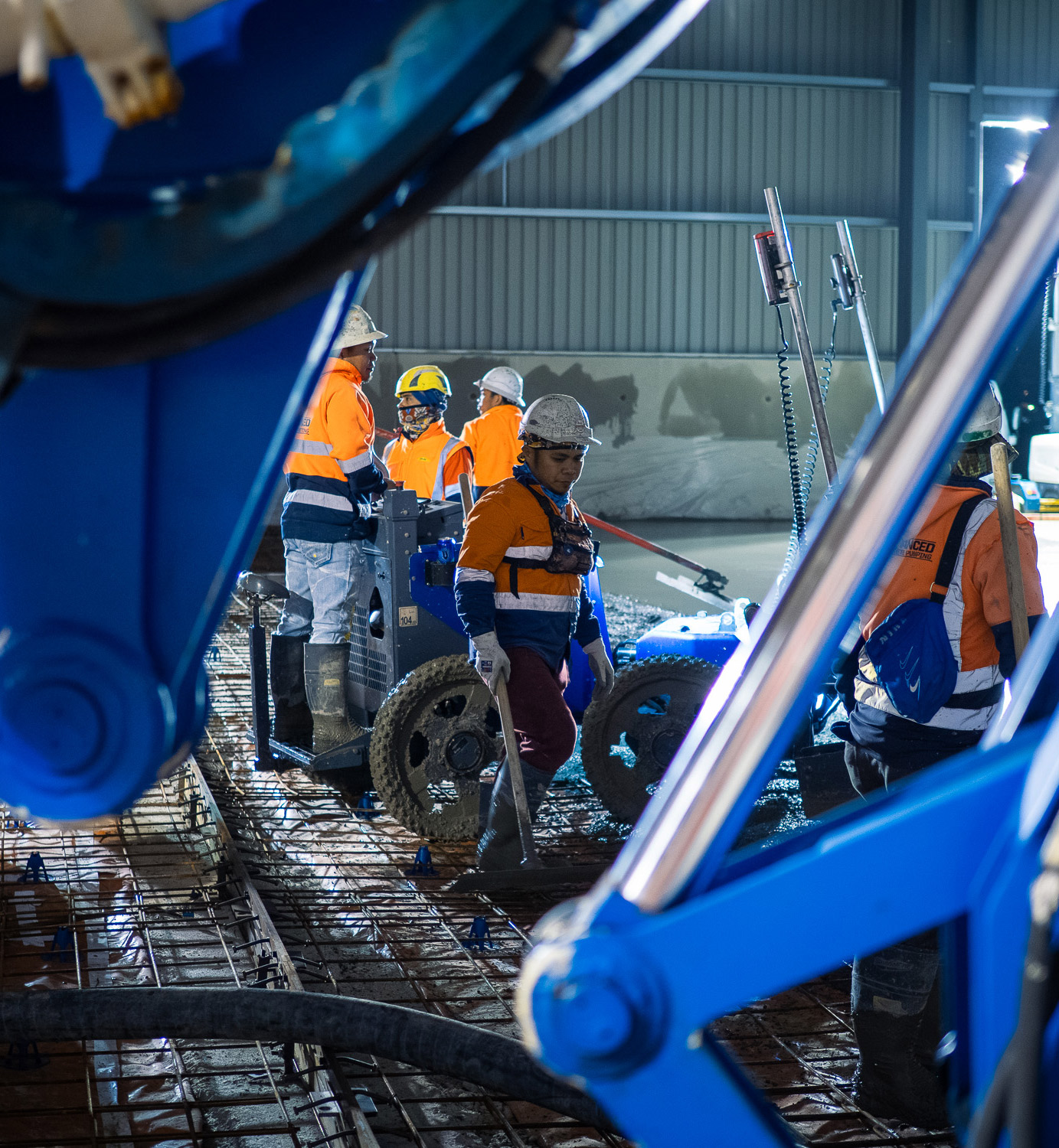 Construction crew in high-visibility jackets and helmets placing concrete inside a large industrial building with reinforcement mesh and pumping equipment.