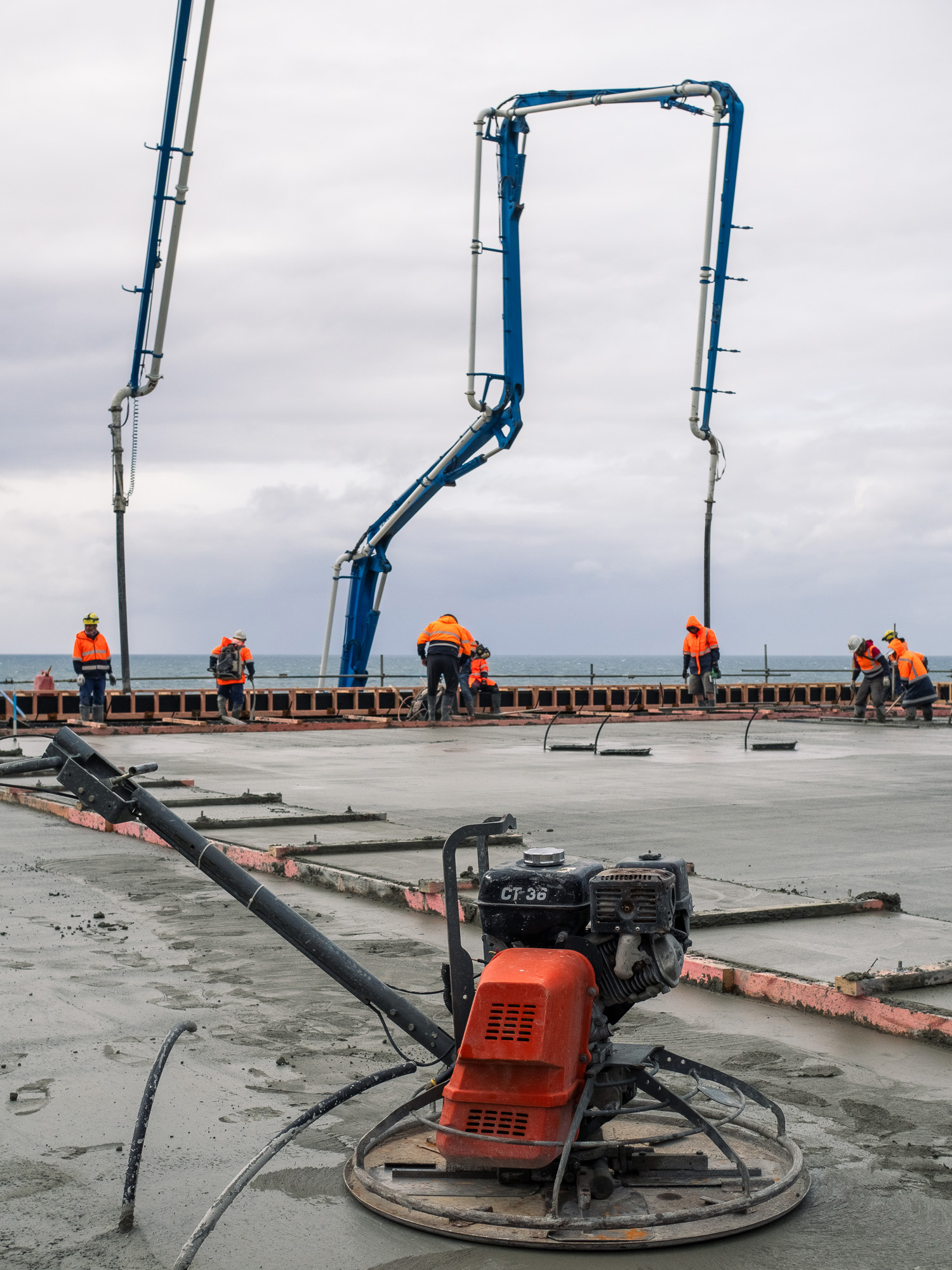 Construction crew in orange high-visibility jackets working with large blue concrete pump booms while pouring and finishing concrete on a coastal site