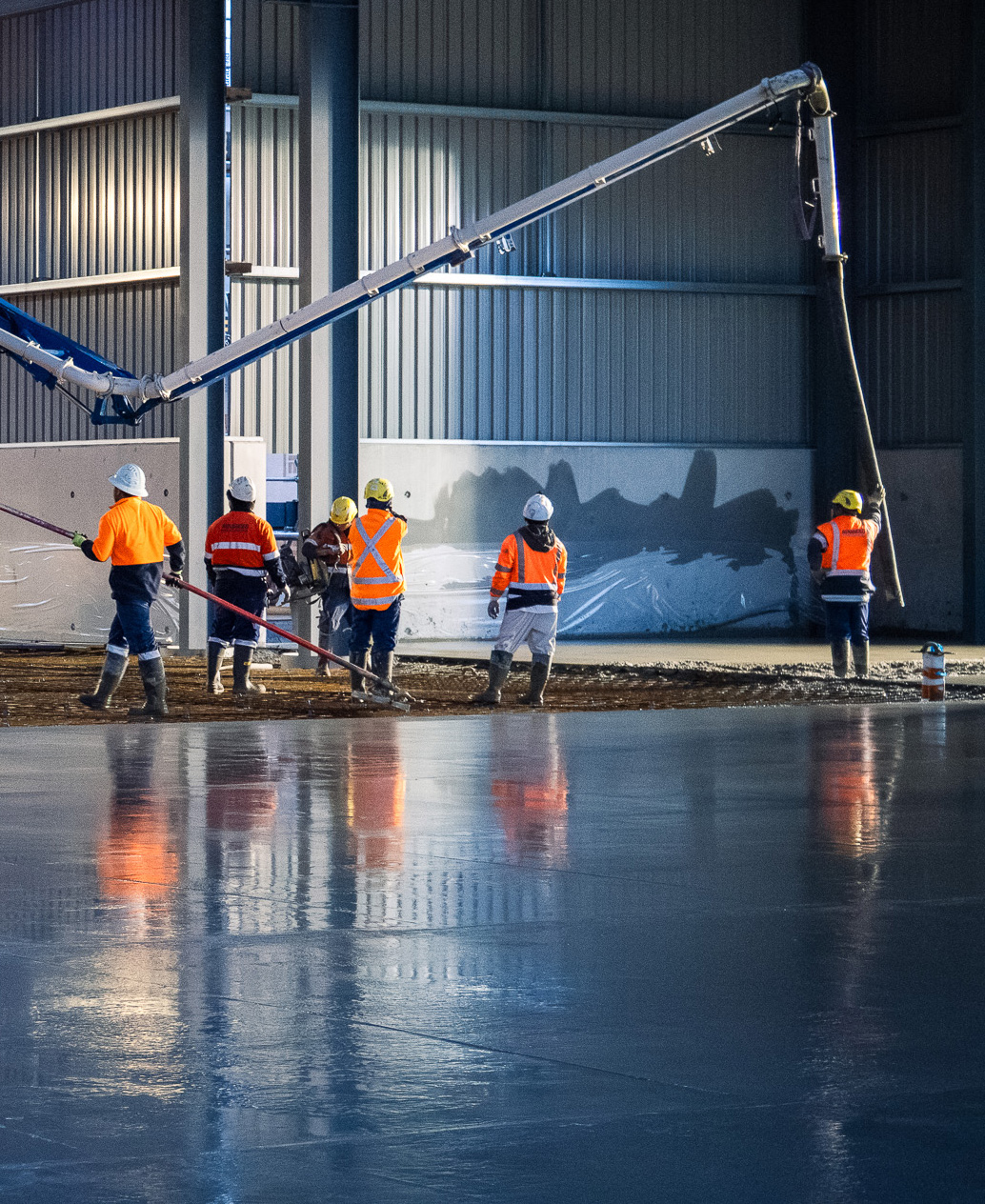 Construction workers in high-visibility jackets and helmets using a concrete pump to pour and level concrete inside a large industrial building