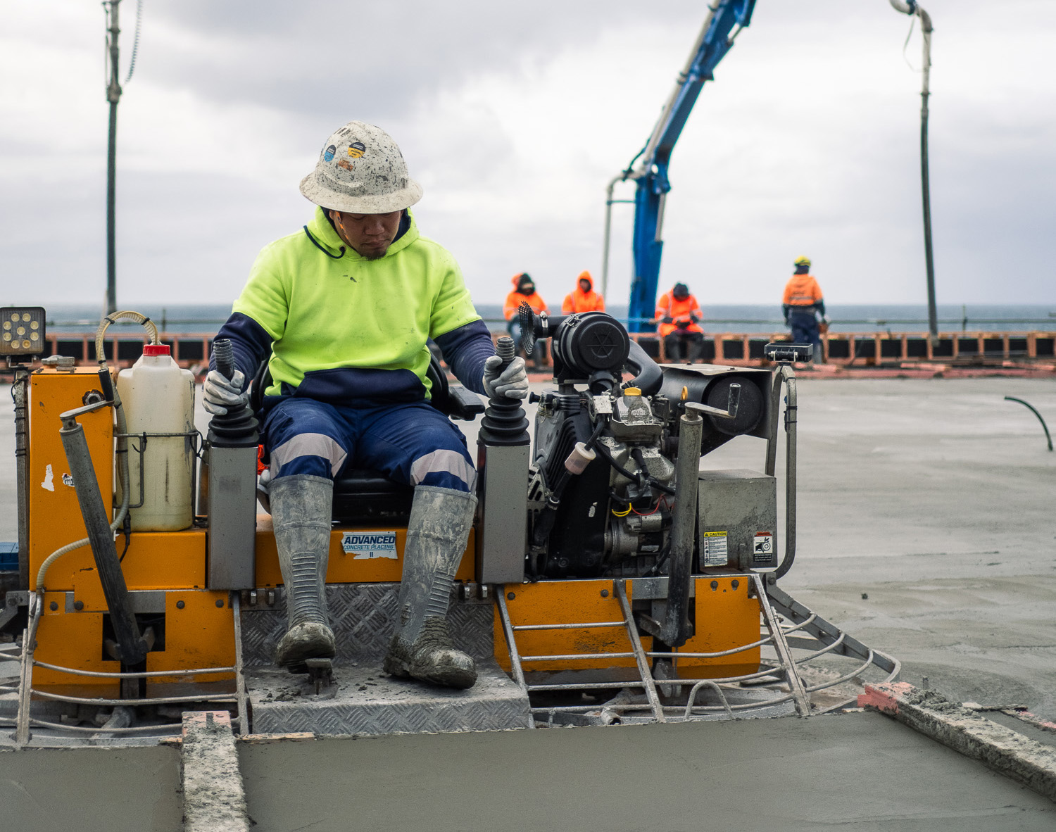 Construction worker in high-visibility gear operating concrete pumping equipment on a coastal building site in New Zealand.