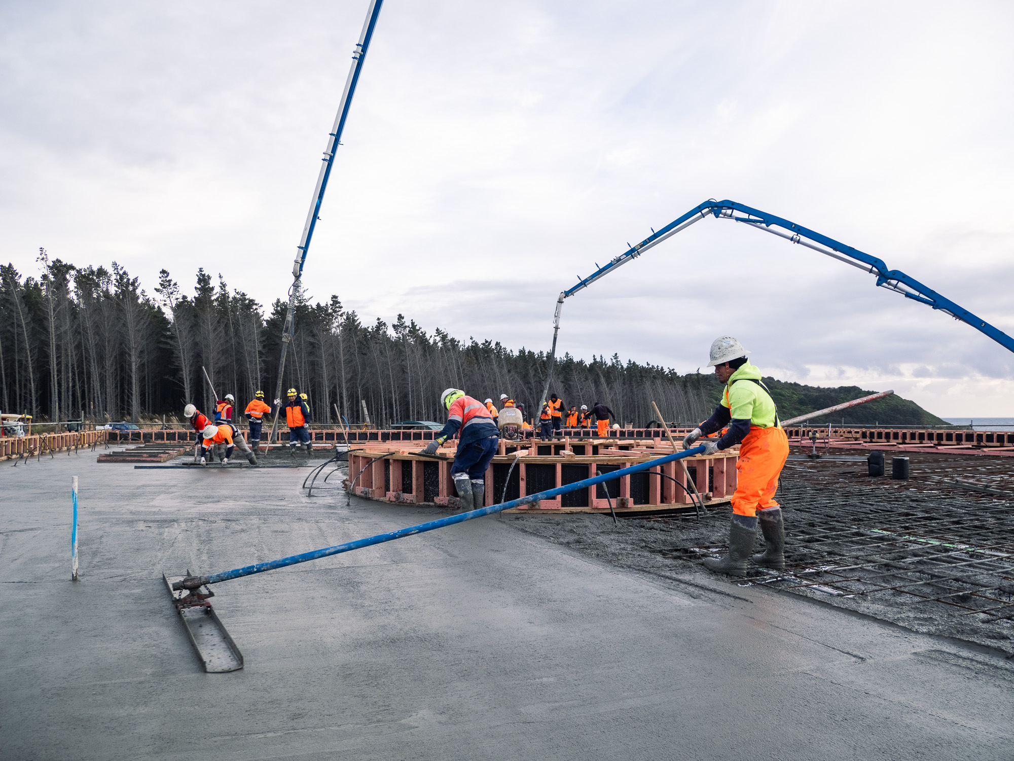 Construction worker in high-visibility gear operating concrete pumping equipment on a coastal building site in New Zealand.