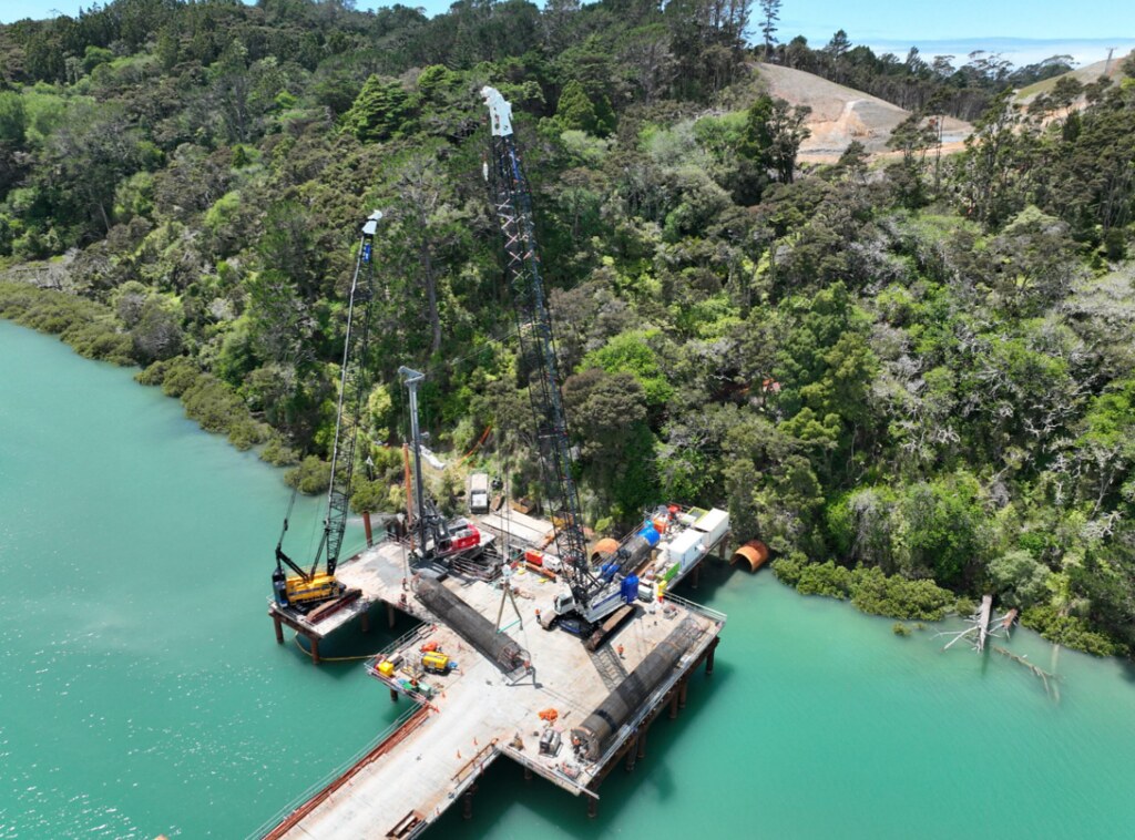 Bridge construction over turquoise water showing cranes, scaffolding, and concrete supports.