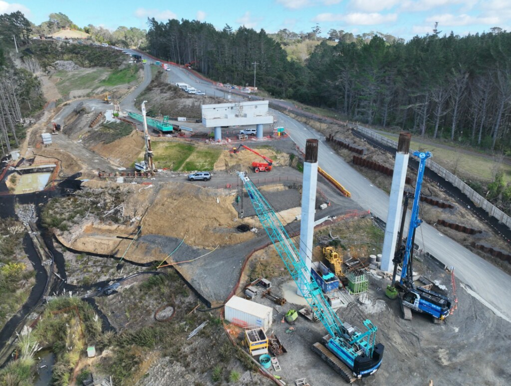 Ground-level construction site with cranes, concrete columns, and earthworks beside a forested area