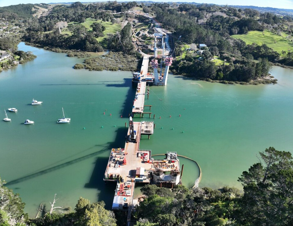 Aerial view of the full bridge span under construction across a river, connecting to roads on both banks.