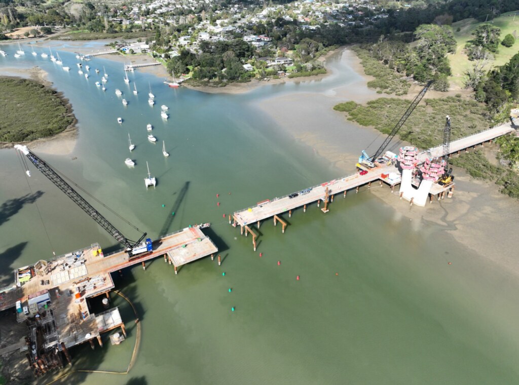 Aerial view of bridge construction across a river estuary with cranes on site and moored boats in the water.
