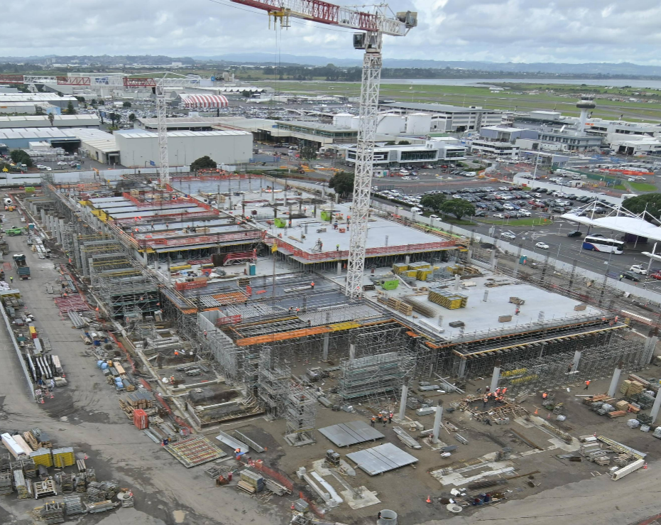 Large construction site with cranes, scaffolding, and concrete slabs being poured near an airport terminal.