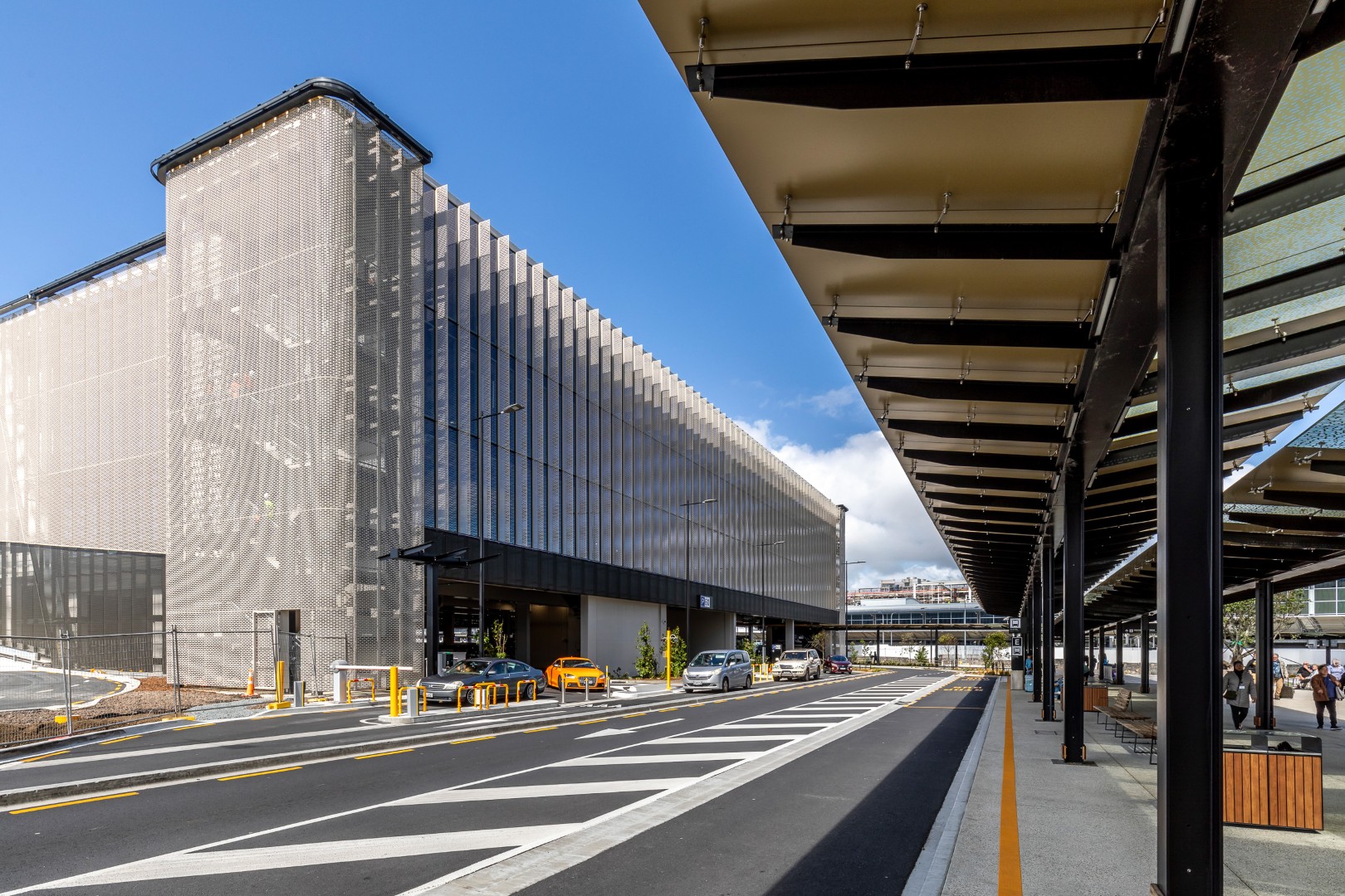 Covered airport pick-up and transport zone with wayfinding signage, buses, cars, and people moving with luggage.