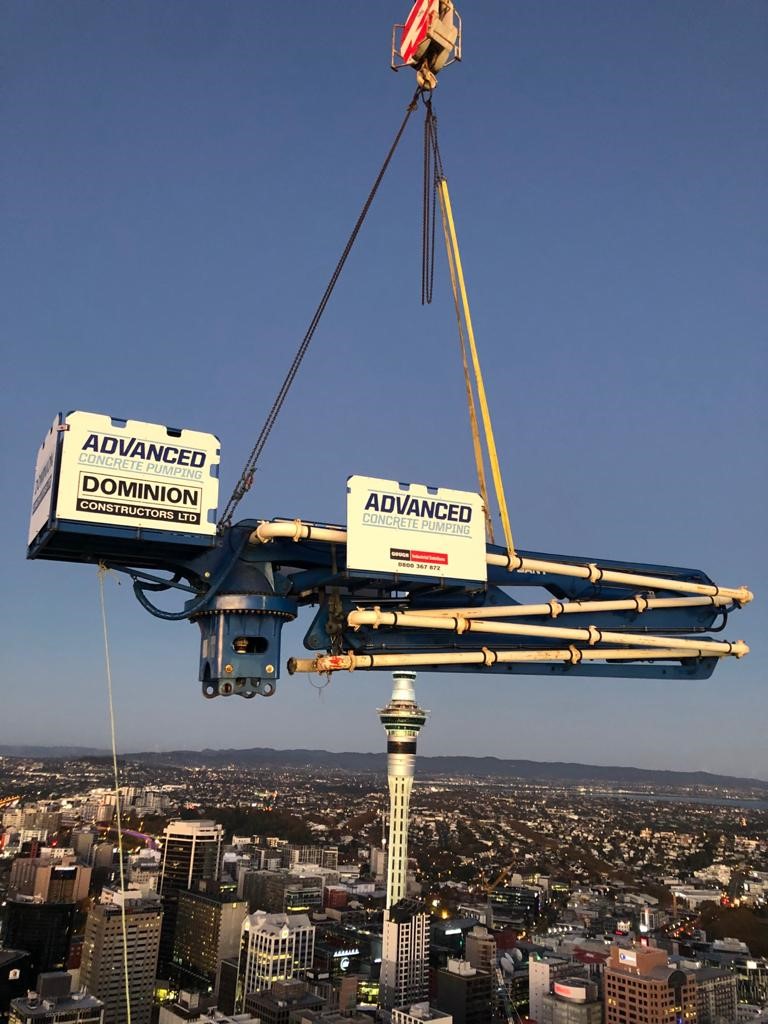 Aerial shot of an Advanced Concrete Pumping boom being lifted by crane high above Auckland city skyline, with the Sky Tower in the background.