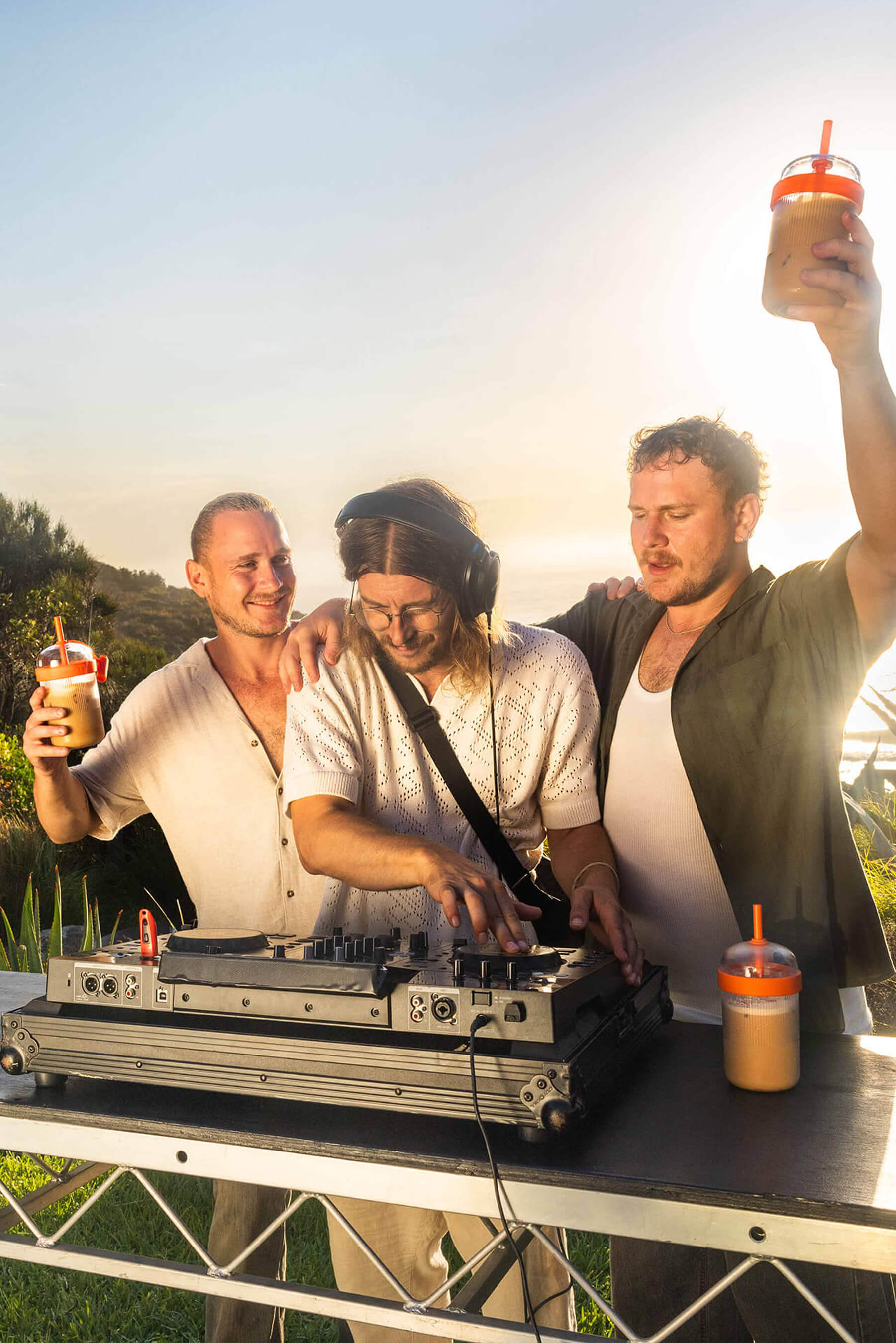 Three friends celebrating outdoors at sunset, holding iced coffee drinks while a DJ plays music for Nespresso’s Happy Hour campaign.