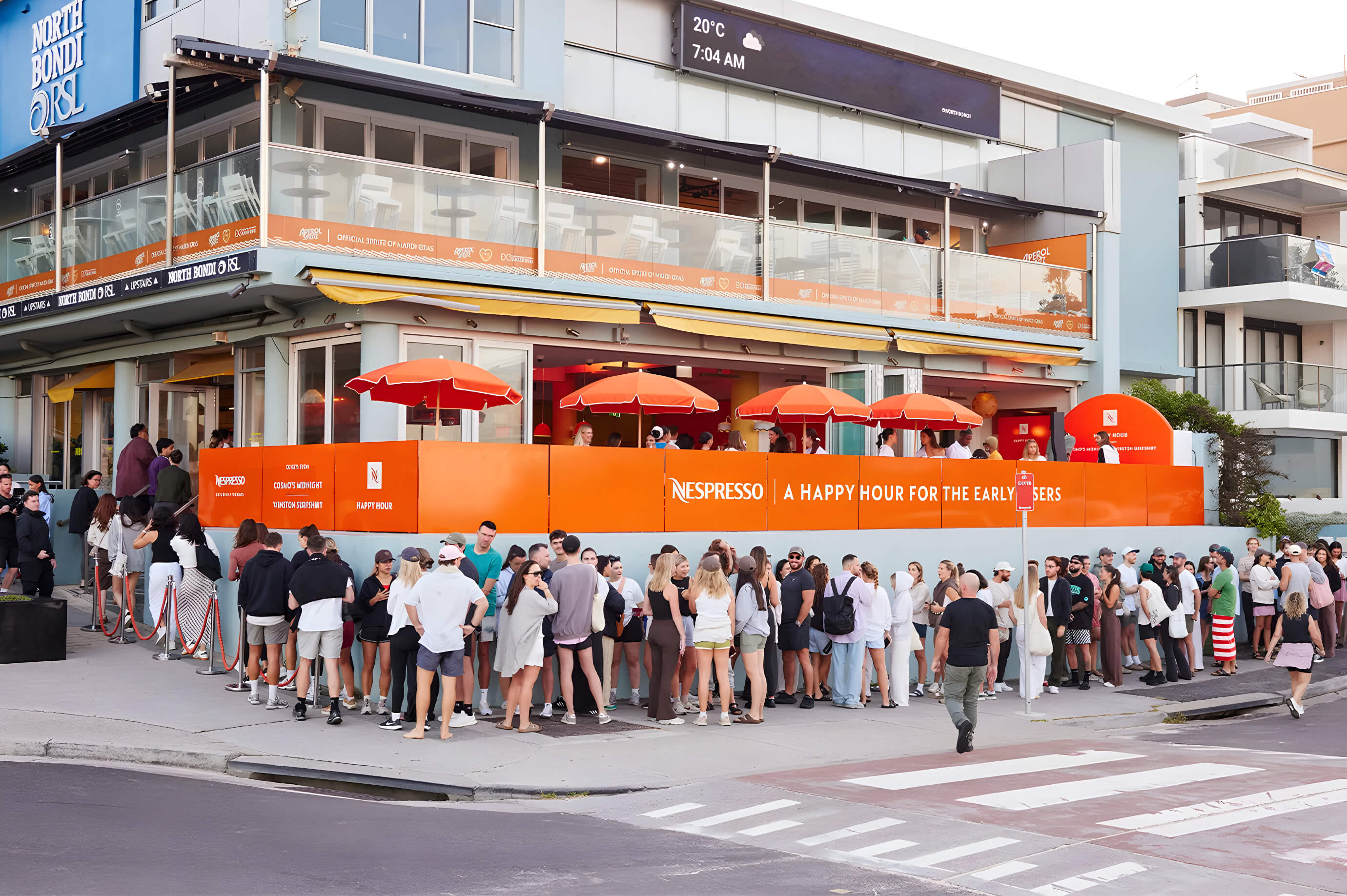 Large crowd queues outside North Bondi RSL for the Nespresso Happy Hour event, with orange umbrellas and branded signage.
