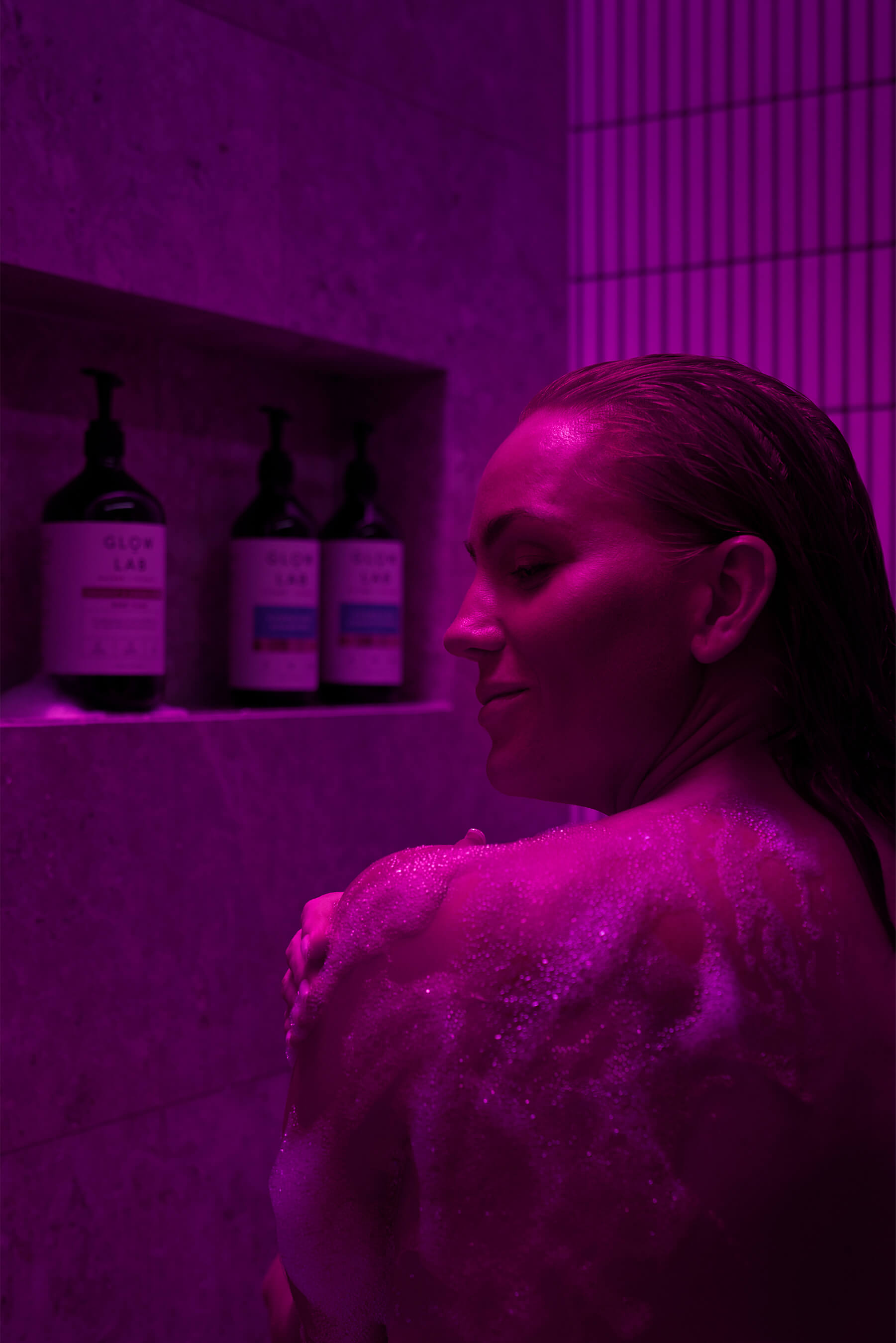 A woman washing her shoulder under deep pink lighting in a stone-tiled shower, with Glow Lab body care bottles softly lit in the niche.