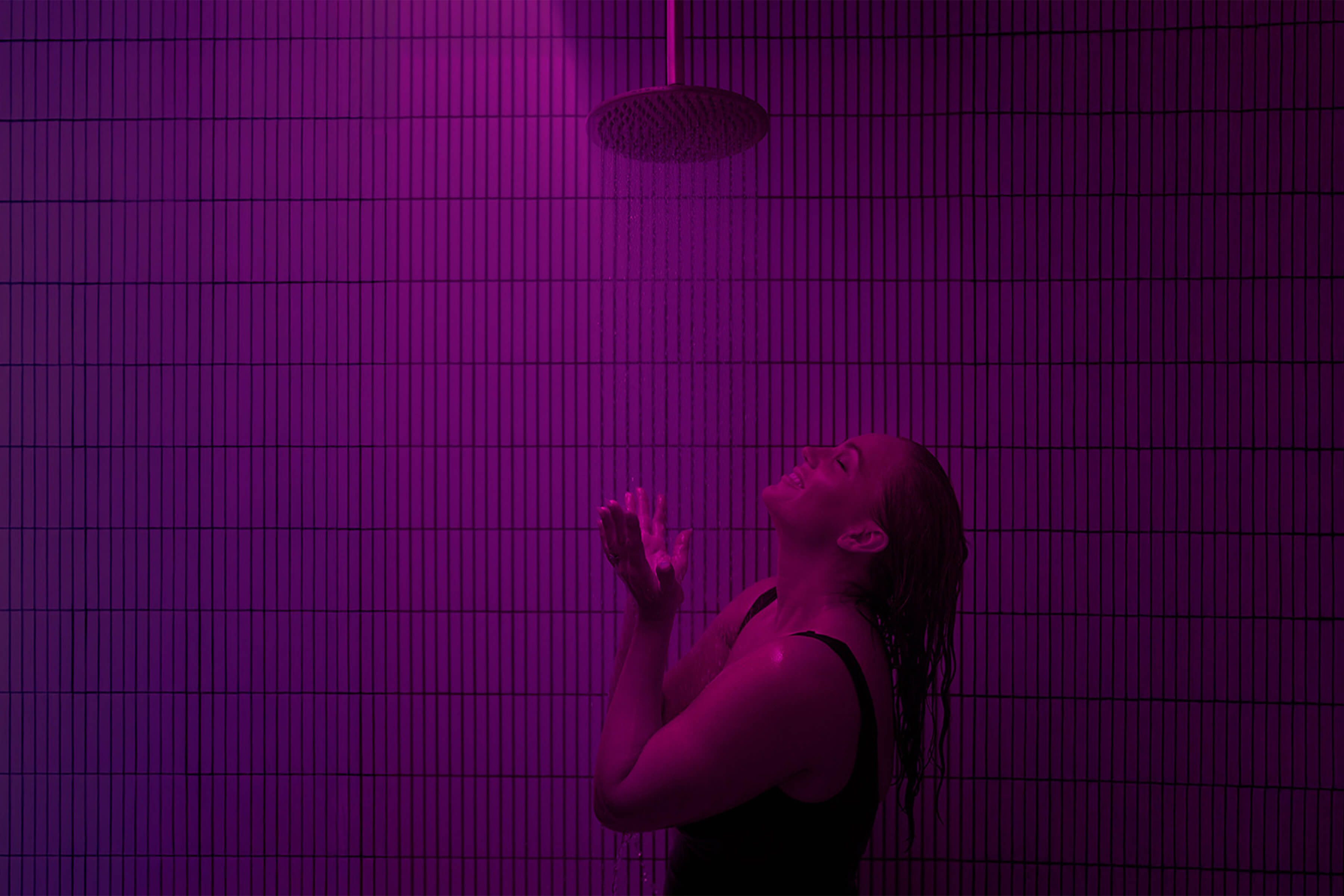 A full-frame image of a woman embracing a rainfall shower, surrounded by vertical white tiles and bathed in Glow Lab’s signature dark pink hue.

