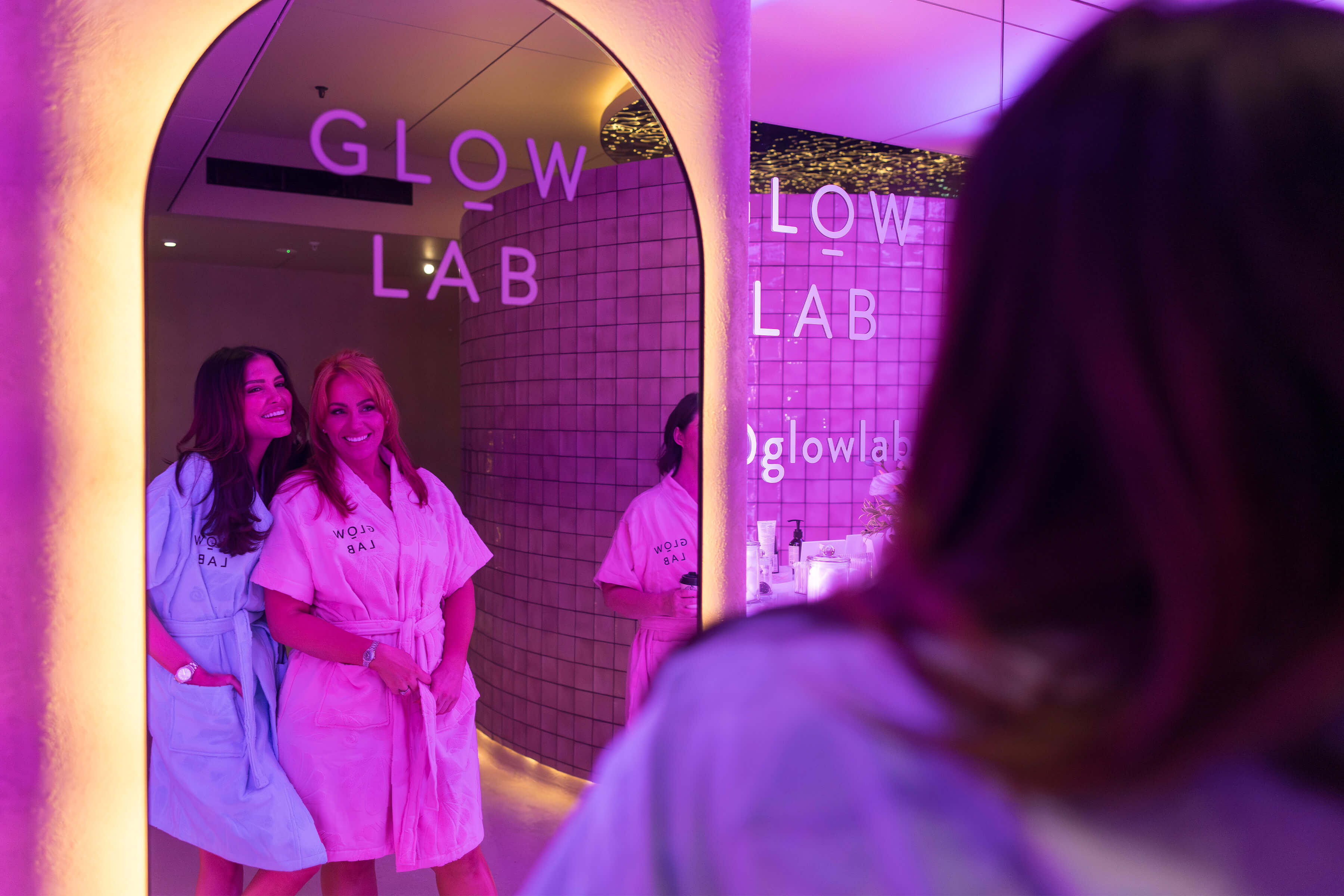 Two women in pastel pink and blue Glow Lab robes smiling into a mirror at the Dark Shower spa, with the Glow Lab logo reflected in glass.