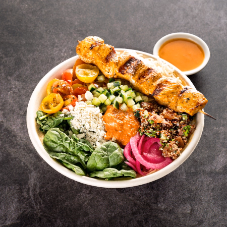 Bowl with grilled chicken kabob over rice, spinach, chopped cucumber, cherry tomatoes, quinoa salad, pickled onions, feta cheese, and red sauce with a small bowl of sauce on the side.