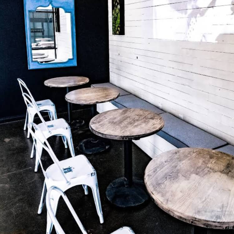 Interior seating area with four round wooden tables, white metal chairs, a long cushioned bench against a white shiplap wall, and a blue framed mirror on a dark wall.