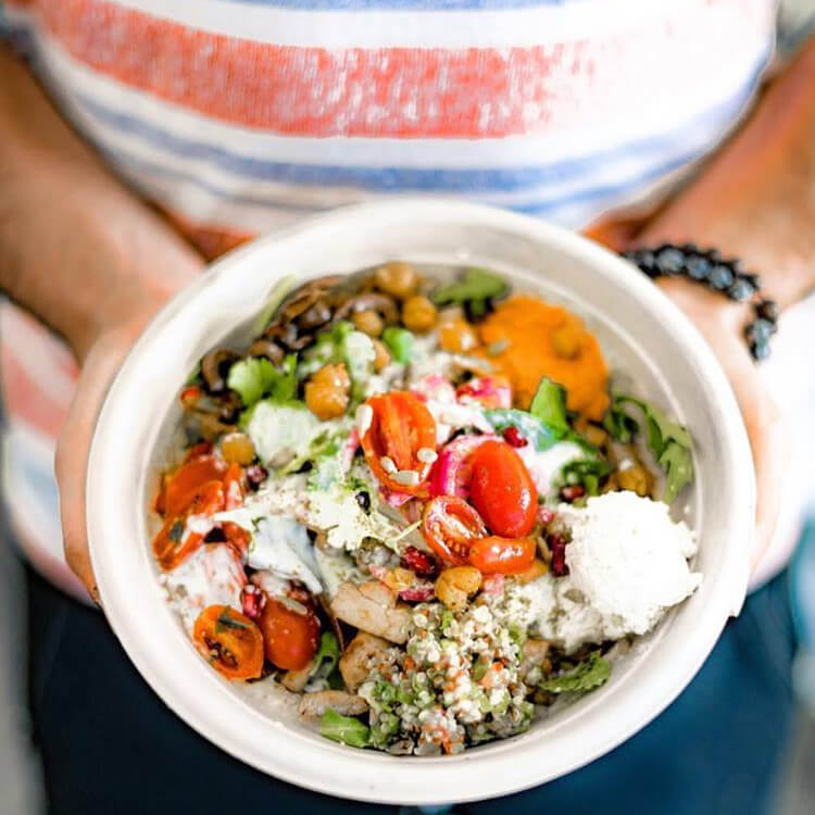 Person holding a healthy bowl with cherry tomatoes, chickpeas, lettuce, grains, and creamy dressing.
