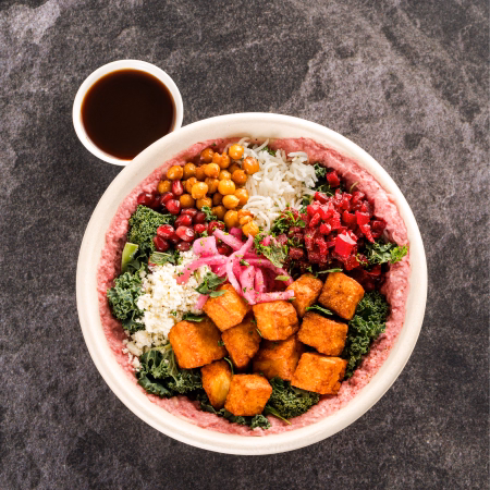 Bowl with kale, fried tofu cubes, pickled onions, feta cheese, chickpeas, pomegranate seeds, rice, and beet salad, with a small cup of dark sauce on the side.