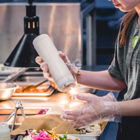 Person wearing plastic gloves squeezes sauce from a white bottle onto food in a kitchen setting.