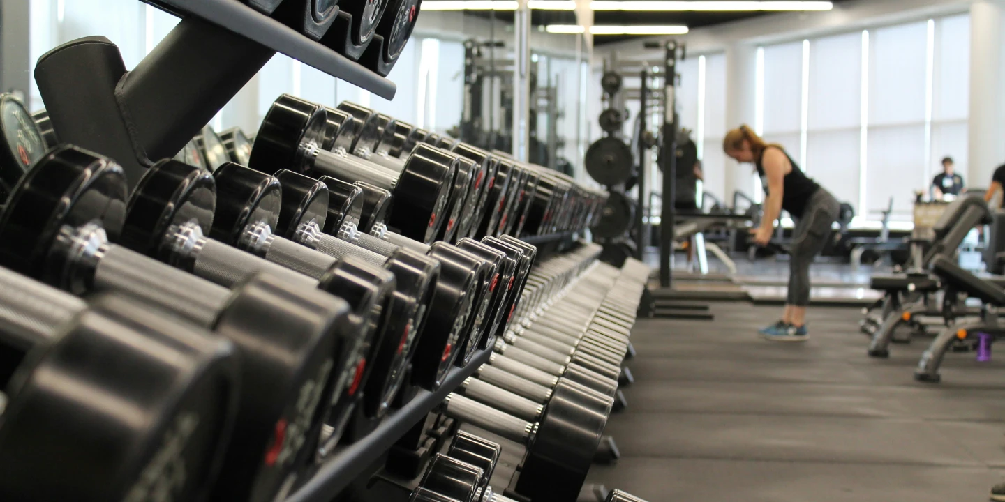 A gym interior at CrossFit Vinita, featuring a row of dumbbells and a woman exercising in the background.