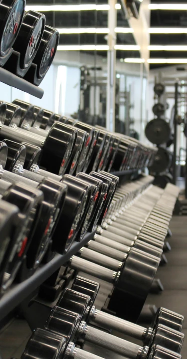 A gym interior at CrossFit Vinita, featuring a row of dumbbells and a woman exercising in the background.