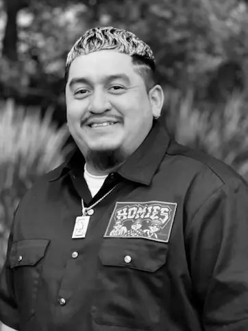 Smiling man with short curly hair and goatee wearing a dark shirt with a 'HOMIES' patch and a pendant necklace.