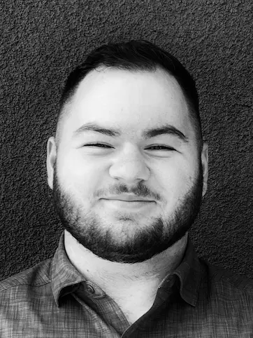 Black and white portrait of a man with short hair and beard smiling with eyes partially closed, wearing a checkered shirt against a textured wall.