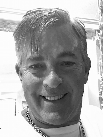 Smiling middle-aged man with short hair wearing a chain necklace indoors near a window with curtains.