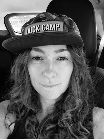 Close-up black-and-white photo of a woman with long wavy hair wearing a camouflage cap with 'DUCK CAMP' on it, sitting inside a car.