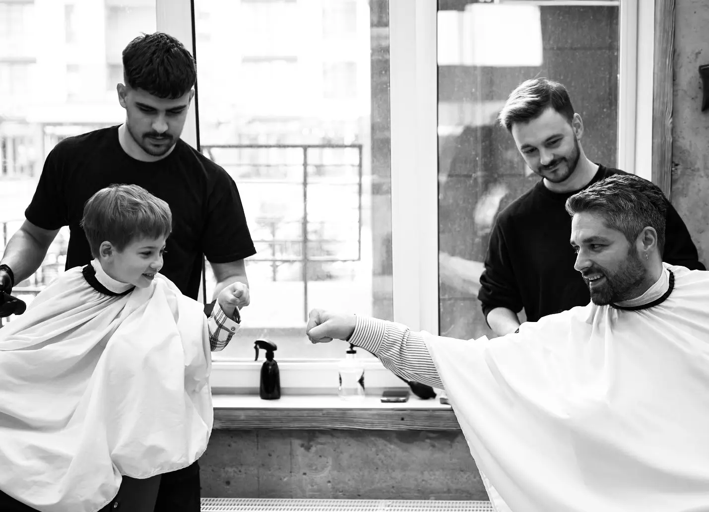 A young boy and an adult man wearing barber capes bump fists while barbers stand behind them in a barbershop.