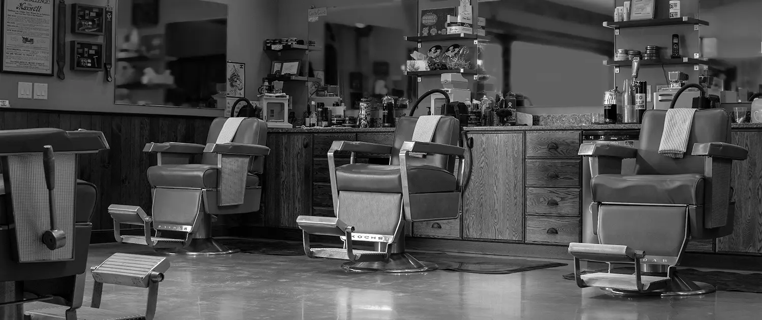 Black-and-white photo of a vintage barbershop interior featuring three classic barber chairs with footrests and towels, wooden cabinets, and large mirrors.