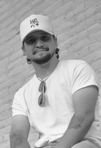 Man with a mustache and goatee wearing a white cap and t-shirt, sitting against a brick wall with sunglasses hanging from his shirt collar.