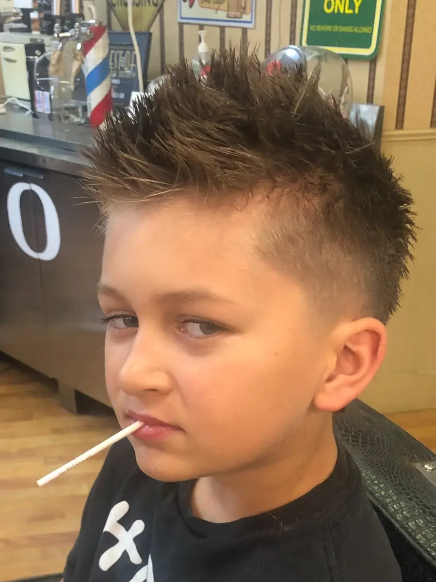 Boy with short spiky brown hair and tapered sides sitting in a barbershop chair with a lollipop stick in his mouth.