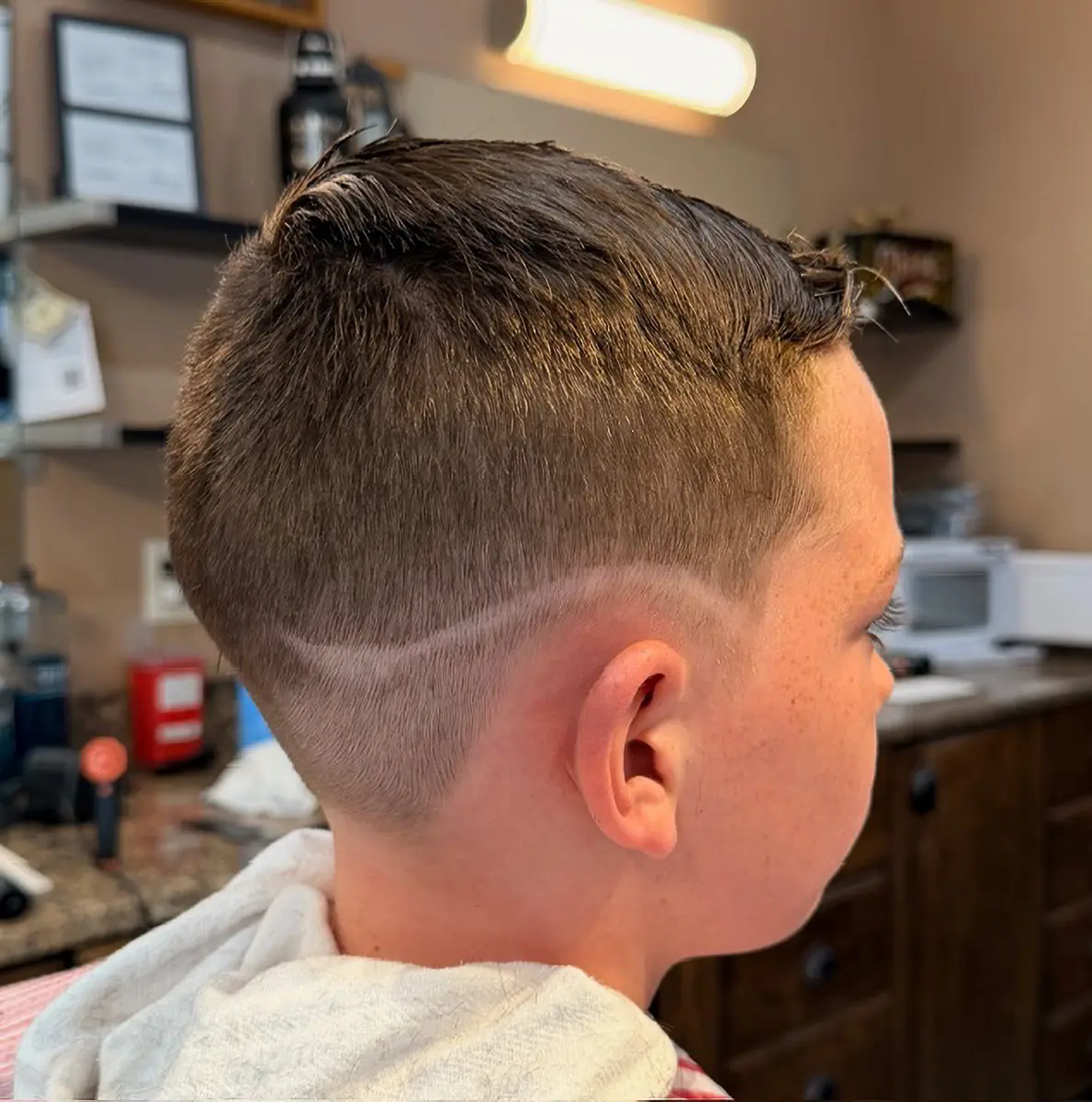 Boy with a short haircut featuring a shaved curved line design on the side of his head in a barbershop.