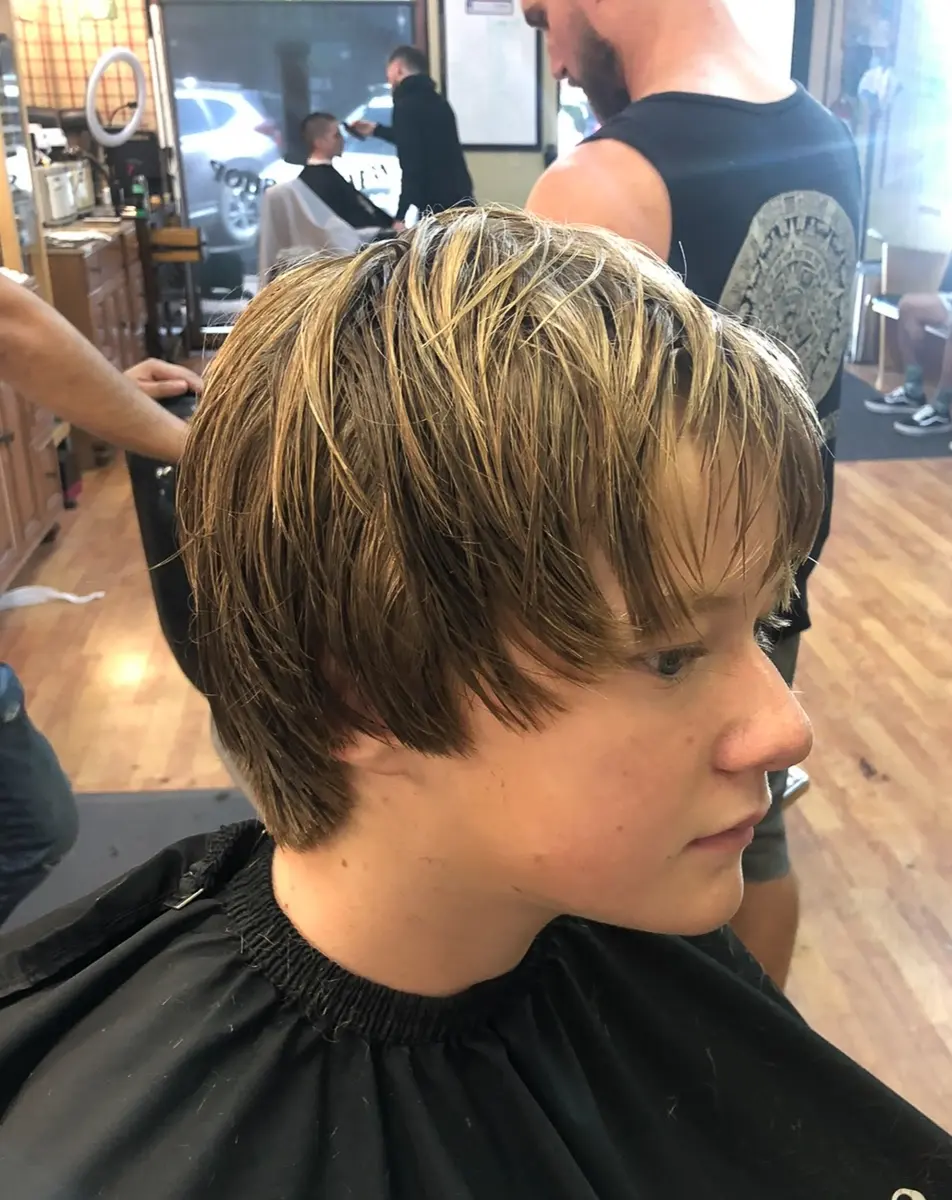 Side profile of a young person with short highlighted hair wearing a black barber cape in a busy barbershop.