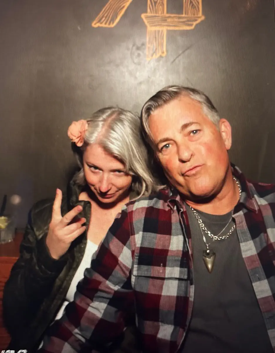 Two adults posing indoors, with the woman on the left flashing a peace sign and the man on the right wearing a plaid shirt and pendant necklace.