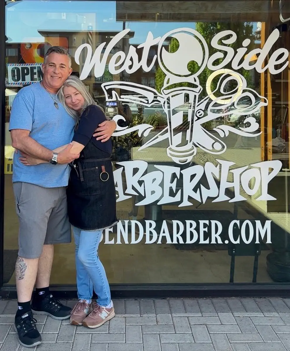 Man in blue shirt and shorts hugging a woman in a black apron and jeans standing outside West Side Barbershop.