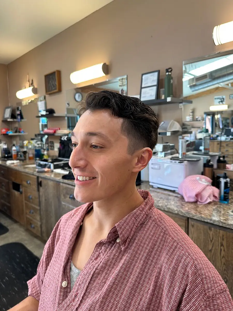 Smiling young man with a fresh haircut wearing a red checkered shirt inside a barbershop.