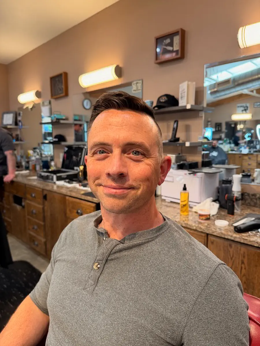 Smiling man with short, styled hair wearing a gray henley shirt sitting in a barbershop with wooden cabinets and grooming products in the background.