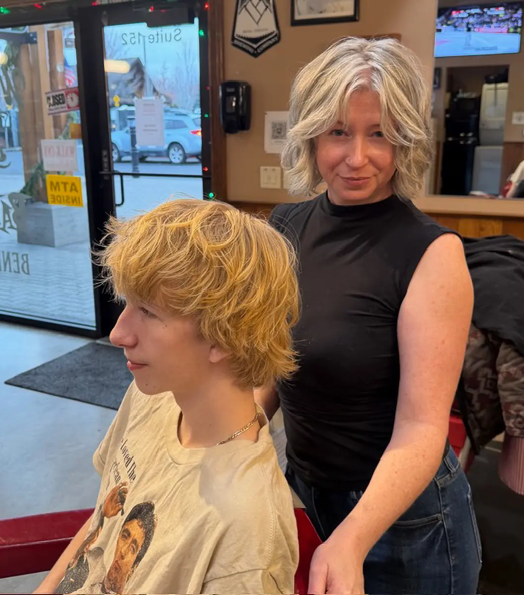 A hairstylist with short layered blonde hair stands behind a seated young man with wavy blonde hair in a salon.