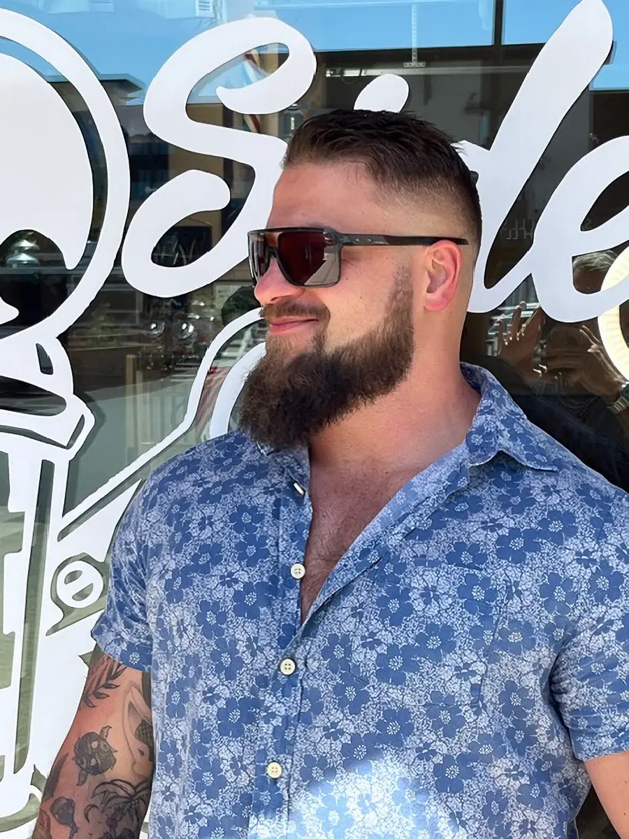 Man with a beard and sunglasses wearing a blue floral shirt standing in front of a glass window with white lettering.