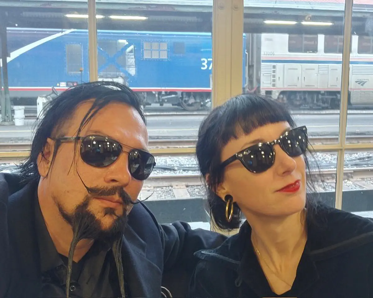 Man and woman wearing sunglasses sitting together at a train station with trains visible outside the window behind them.