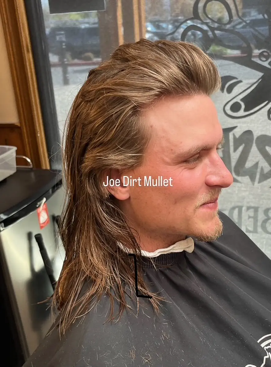 Man with a styled Joe Dirt mullet haircut sitting in a barber chair inside a salon.